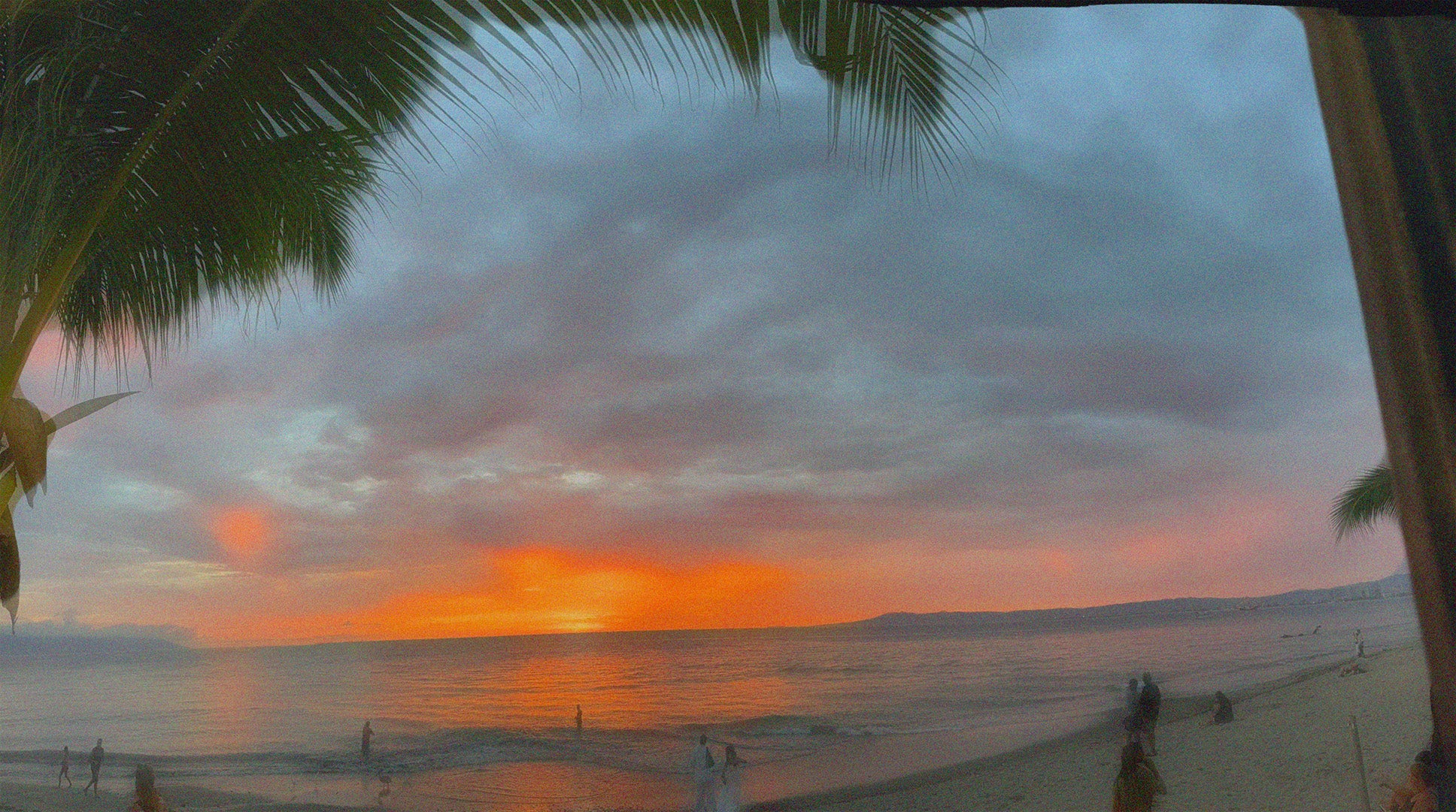 Beach sunset with glowing orange and clouded sky, palm leaves framing the scene, and people in the distance by the ocean and on the sand.
